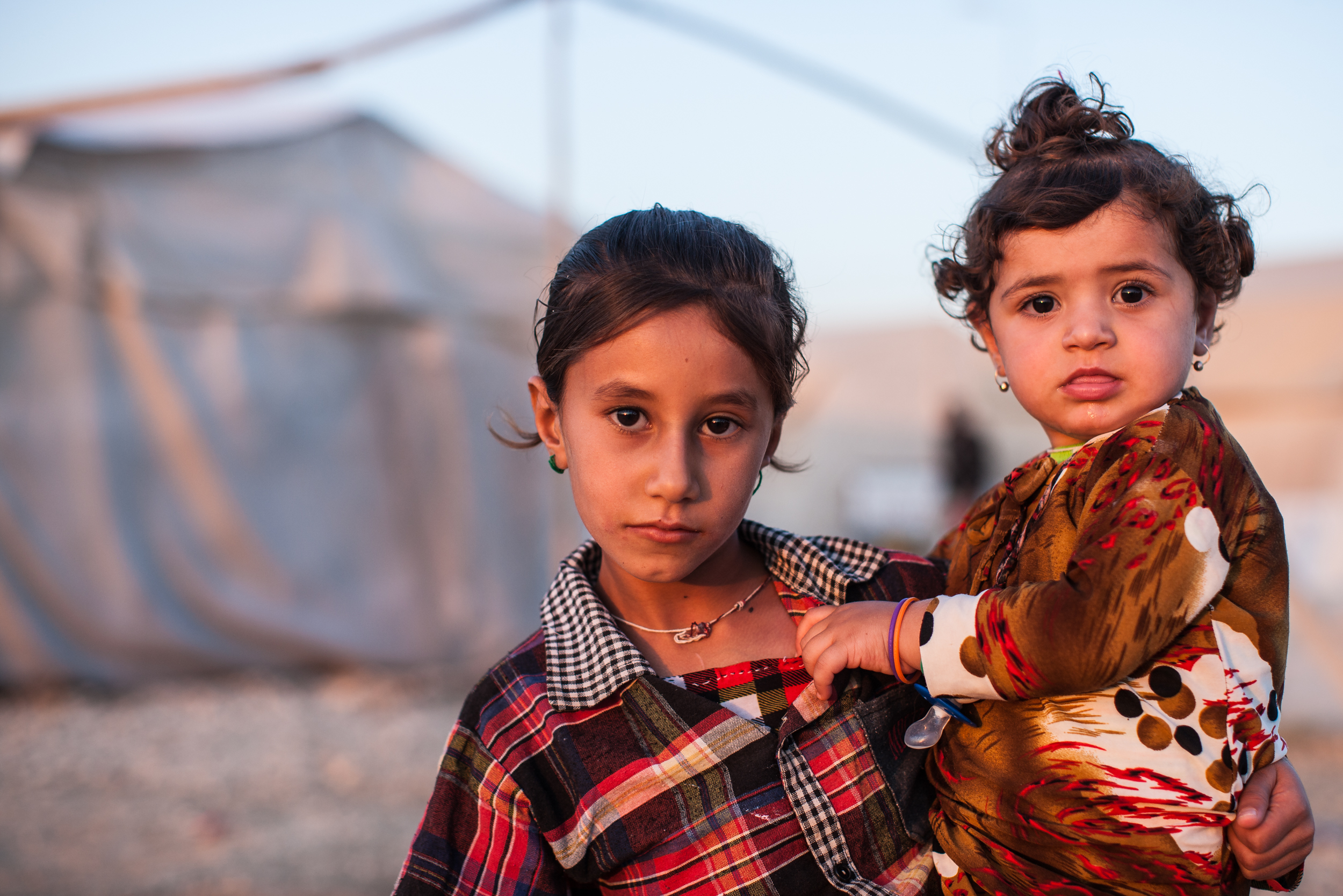 A young Yezidi girl carries her sister in a displaced persons camp in Dohuk, Iraq.