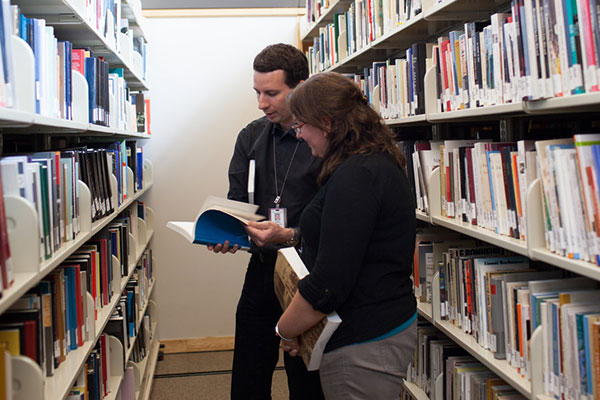 Two people read a book between stacks at the Museum Library