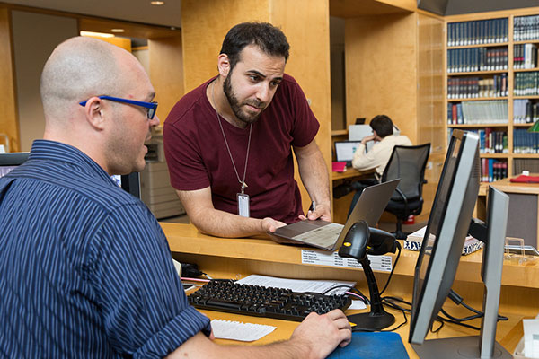 A Museum librarian sits at a computer and discusses a question with a Museum visitor